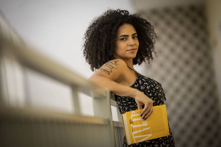 A imagem mostra uma mulher com cabelo cacheado e volumoso， usando um vestido preto com estampas florais. Ela está encostada em uma grade， segurando um livro amarelo. O fundo é desfocado， mas sugere um ambiente interno com luz natural