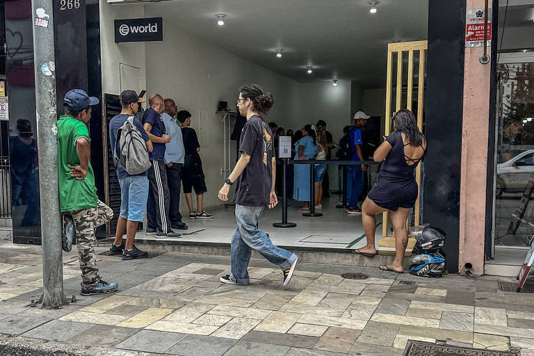 A imagem mostra uma rua movimentada com várias pessoas em frente a um edifício. Algumas pessoas estão entrando e saindo do local， enquanto outras estão paradas conversando. O ambiente parece urbano， com calçadas e um piso de pedras. Há uma mistura de roupas， incluindo uma pessoa vestindo uma camiseta verde. O céu está nublado.