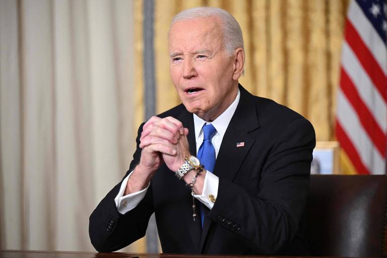 Biden de terno escuro e gravata azul está sentado em uma cadeira， com as mãos unidas em frente ao corpo， parecendo expressar preocupação ou emoção. Ao fundo， há cortinas douradas e uma bandeira dos Estados Unidos visível.