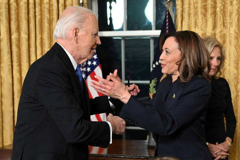 A imagem mostra Joe Biden e Kamala Harris se cumprimentando em um ambiente formal， no Salão Oval. Ao fundo， há cortinas douradas e bandeiras dos Estados Unidos. Uma terceira pessoa está visível ao fundo， observando a interação.