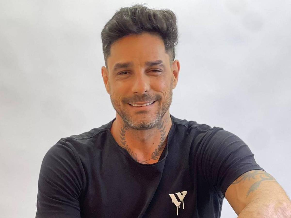 Diego Grossi
 *** Local Caption *** Um homem está sentado em uma cadeira, sorrindo para a câmera. Ele usa uma camiseta preta com um logotipo pequeno no peito e calças pretas rasgadas. Seus braços e mãos estão cobertos de tatuagens, e ele usa um relógio preto no pulso. O fundo é claro e desfocado.