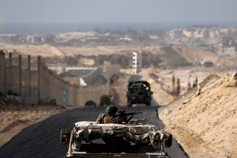 A imagem mostra um caminho em uma área de conflito， com veículos militares em movimento. No primeiro plano， um veículo está parcialmente coberto， com um soldado visível. Ao fundo， há uma paisagem que inclui uma barreira e uma vista do mar. O céu está claro e a área ao redor parece árida