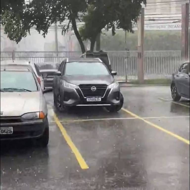 A imagem mostra um estacionamento durante uma forte chuva. Um carro preto está estacionado em uma das vagas， enquanto outros carros estão visíveis ao fundo. A chuva é intensa， e a água está acumulada no chão， refletindo a luz. Árvores estão ao redor， e o ambiente parece nublado.