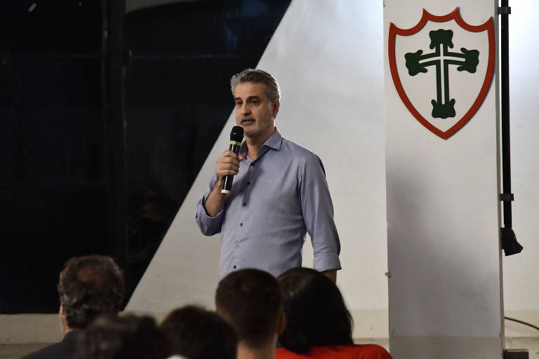 Um homem está em pé， falando ao microfone em um evento. Ele usa uma camisa de manga longa azul clara e tem cabelo grisalho. Ao fundo， há uma parede com um escudo que apresenta uma cruz e um contorno vermelho. A plateia é visível， com pessoas sentadas， mas seus rostos não estão claramente visíveis.