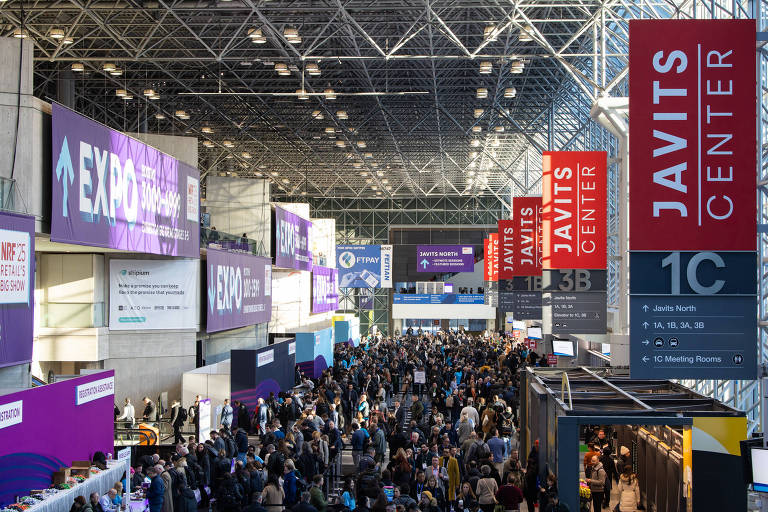 A imagem mostra o interior do Centro de Convenções Javits, com várias pessoas se movendo entre estandes. Há grandes banners suspensos com as palavras 'JAVITS CENTER' e 'EXPO', além de outros sinais informativos. O ambiente é amplo e iluminado, com uma estrutura de teto de vidro.