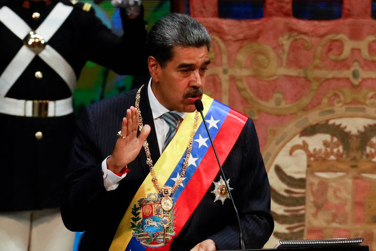 Maduro em um terno escuro está falando ao microfone， levantando a mão direita em um gesto de saudação. Ele usa uma faixa tricolor (amarela， azul e vermelha) e um colar de medalhas. Ao fundo， há uma decoração elaborada e uma figura em uniforme militar.