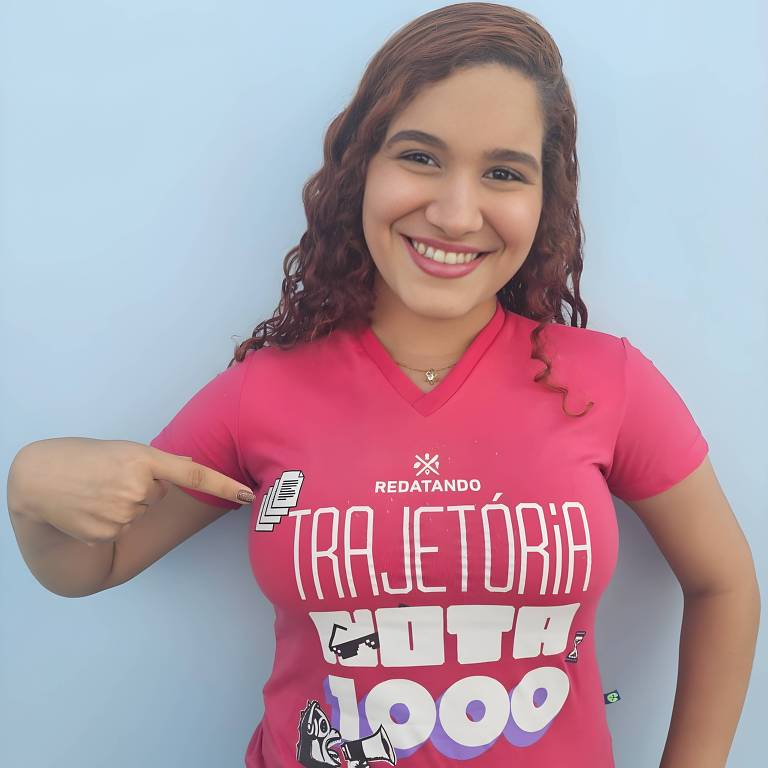 Uma pessoa sorridente está usando uma camiseta rosa com a palavra 039;TRAJETÓRIA039; em destaque， acompanhada de outros elementos gráficos. Ela está apontando para a camiseta com uma mão e com a outra mão na cintura. O fundo é de uma parede azul.