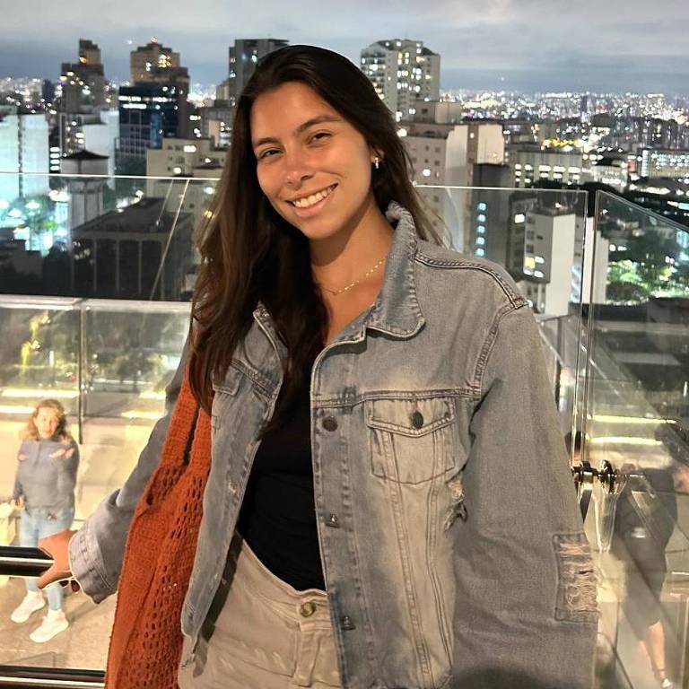 Uma jovem mulher está em primeiro plano， sorrindo， com um casaco jeans e uma blusa preta. Ela segura uma bolsa laranja. Ao fundo， há uma vista noturna da cidade， com prédios iluminados e um céu parcialmente nublado.