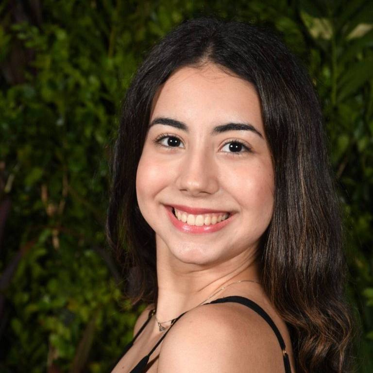 A imagem mostra uma jovem mulher sorrindo， com cabelo longo e ondulado. Ela está usando uma blusa preta e está posicionada de lado， com um fundo de folhagens verdes.