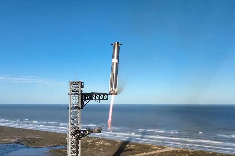 A imagem mostra uma estrutura de lançamento de foguete em uma plataforma， com o mar ao fundo e um céu azul claro. A estrutura é metálica e se eleva em direção ao céu， enquanto as ondas do mar são visíveis na parte inferior da imagem. O foguete se aproxima da plataforma