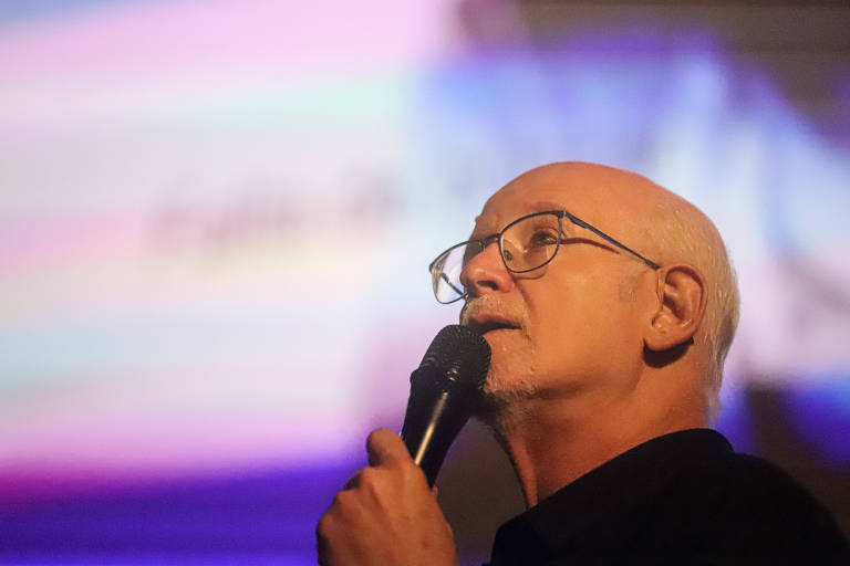 Um homem de cabelos brancos e calvo, usando óculos, está falando ao microfone. Ele parece estar em um evento ou apresentação, com um fundo desfocado que apresenta cores suaves, como rosa e azul.