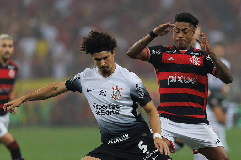 A imagem mostra um momento de uma partida de futebol， onde um jogador com a camisa branca e preta está prestes a chutar a bola. Ele é cercado por dois jogadores adversários， um deles vestindo uma camisa vermelha e preta. O fundo apresenta uma multidão de torcedores， e o clima parece ser de grande expectativa.
