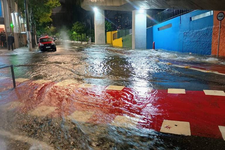 A imagem mostra uma rua inundada durante a noite， com água cobrindo a maior parte do asfalto. Há um carro vermelho estacionado à esquerda e luzes de postes iluminando a cena. Ao fundo， há uma parede azul e uma estrutura elevada， possivelmente um viaduto. A água parece estar em movimento， criando redemoinhos na superfície.