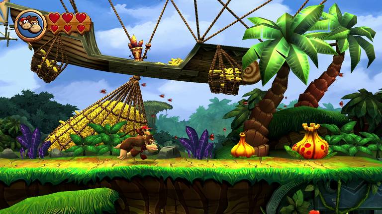 A imagem mostra uma cena de um jogo de plataforma, onde um personagem semelhante a um macaco, chamado Donkey Kong, está correndo em um cenário tropical. Ao fundo, há uma estrutura de madeira suspensa com cestas cheias de bananas. O ambiente é repleto de palmeiras, plantas coloridas e um céu azul com nuvens. No canto superior esquerdo, há um ícone com rostos de personagens e corações, indicando a vida do jogador.