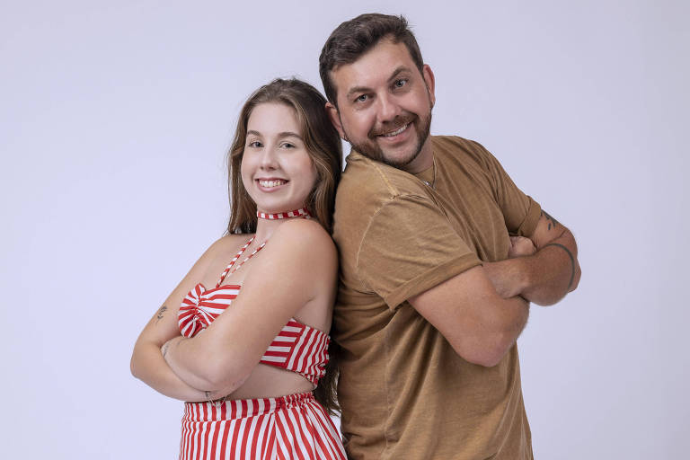  A imagem mostra um casal posando de costas um para o outro. A mulher usa um conjunto de top e saia longa listrada em vermelho e branco, com uma fenda na saia. Ela tem cabelo longo e solto, e está sorrindo. O homem está vestindo uma camiseta marrom e jeans, com um sorriso no rosto. Ambos estão com os braços cruzados e a imagem tem um fundo branco