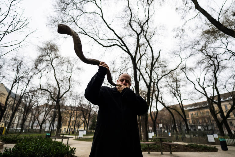 Um homem está tocando um shofar, um instrumento musical feito de chifre, em um parque. Ele está vestido com roupas escuras e se posiciona em meio a árvores sem folhas, sugerindo um clima de outono ou inverno. O fundo apresenta edifícios e um espaço verde.