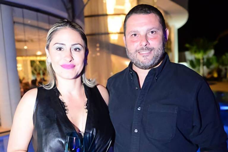 A imagem mostra um casal posando juntos em um evento social. A mulher está usando um vestido preto e segurando um copo， enquanto o homem está vestido com uma camisa preta. Ao fundo， há uma iluminação suave e uma estrutura arquitetônica visível.