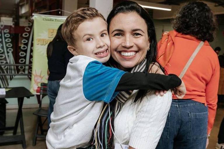 A imagem mostra uma mulher sorridente segurando uma criança que a abraça de forma afetuosa. A criança， também sorridente， veste uma camisa branca com detalhes azuis. O ambiente parece ser um local público， com outras pessoas e elementos ao fundo， como mesas e objetos decorativos. A cena transmite alegria， carinho e conexão entre os dois.