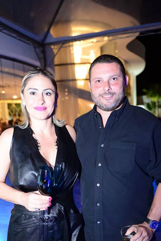 A imagem mostra um casal posando juntos em um evento social. A mulher está usando um vestido preto e segurando um copo, enquanto o homem está vestido com uma camisa preta. Ao fundo, há uma iluminação suave e uma estrutura arquitetônica visível.