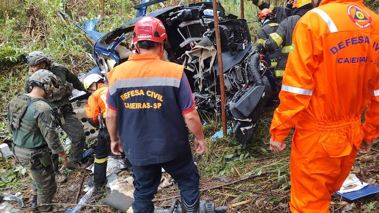 A imagem mostra um acidente de veículo em uma área de vegetação densa. Várias pessoas， incluindo membros da Defesa Civil e bombeiros， estão ao redor de um carro azul que está severamente danificado. Os socorristas estão usando equipamentos de resgate e vestindo uniformes de emergência. O ambiente é de floresta， com árvores e vegetação ao redor.