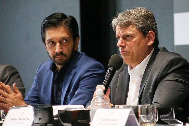 A imagem mostra dois homens em uma mesa de conferência. O homem à esquerda， com cabelo escuro e barba， usa uma camisa azul e parece estar ouvindo atentamente. O homem à direita， com cabelo grisalho e usando um terno escuro， está falando ao microfone. Há copos e garrafas de água sobre a mesa， além de um nome visível em uma placa na frente deles.
