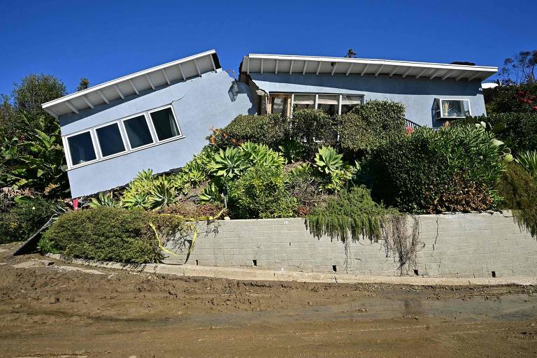 A imagem mostra uma casa de cor azul inclinada para o lado， com um jardim ao redor. A estrutura parece ter se deslocado， com a parte inferior da casa visivelmente mais alta que a parte superior. O céu está limpo e azul， e há vegetação densa ao redor da casa.