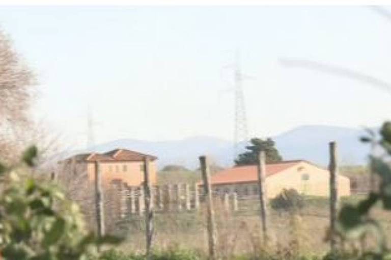 A imagem mostra uma paisagem rural em Roma， com um campo e algumas construções ao fundo. Há árvores e uma cerca visíveis， além de uma torre de comunicação