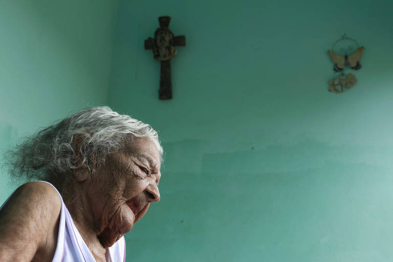 A imagem mostra uma mulher idosa com cabelo grisalho, sentada e olhando para baixo. Ela está vestindo uma blusa branca e o fundo é de uma parede verde clara. No fundo, há uma cruz na parede e uma borboleta decorativa.