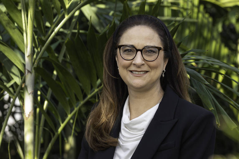 Uma mulher sorridente está em pé em frente a um fundo de plantas verdes. Ela usa óculos e está vestida com uma blusa clara e um blazer escuro. O ambiente parece ser ao ar livre， com folhagens ao redor.