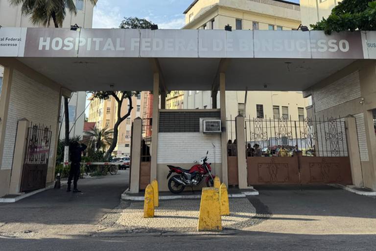 Iimagem mostra a entrada de um hospital， com um grande portão e uma placa na parte superior. O ambiente é urbano， com edifícios ao fundo e algumas árvores visíveis. Há uma motocicleta estacionada à esquerda e pessoas próximas ao portão. O céu está claro.