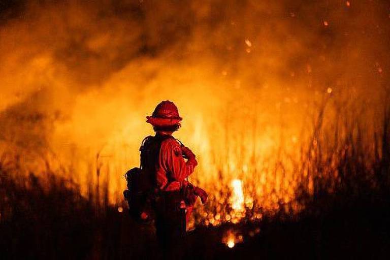 A imagem mostra um bombeiro de costas， usando um capacete e uma mochila， observando um incêndio florestal. Chamas intensas e fumaça densa iluminam a cena， criando um ambiente dramático e perigoso.