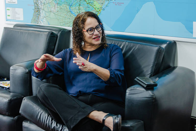 Uma mulher sentada em um sofá preto， usando uma blusa azul e óculos. Ela está gesticulando com as mãos enquanto conversa. Ao fundo， há um mapa na parede.
