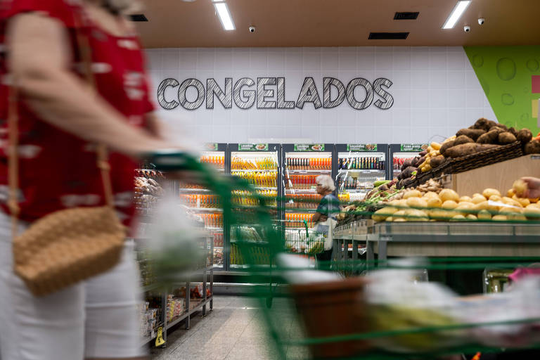 A imagem mostra uma seção de congelados em um supermercado. Ao fundo， há um grande letreiro escrito 039;CONGELADOS039;. Na frente， uma pessoa empurra um carrinho de compras， enquanto prateleiras com produtos congelados e frutas estão visíveis. A iluminação é clara e o ambiente é organizado.