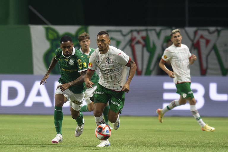 A imagem mostra uma cena de uma partida de futebol， onde um jogador com uniforme verde está correndo atrás de um jogador com uniforme branco e detalhes em verde. O jogador em branco está driblando a bola， enquanto outros jogadores estão ao fundo， observando a jogada. O campo é bem iluminado e há uma bandeira ao fundo com um desenho que parece ser de um animal.