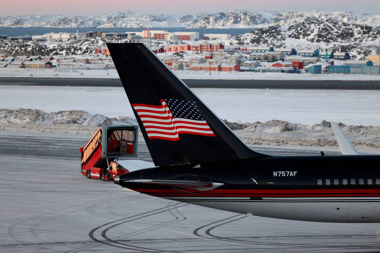 A imagem mostra a parte traseira de um avião estacionado em um aeroporto coberto de neve. O avião tem uma cauda com um design que inclui as cores da bandeira dos Estados Unidos. Ao fundo， é possível ver uma área urbana com edifícios e montanhas cobertas de neve.