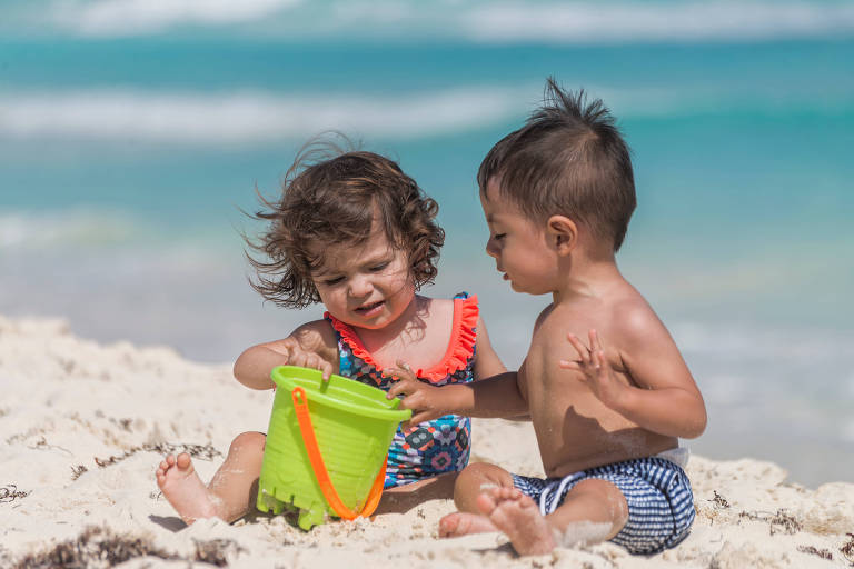 Duas crianças pequenas estão sentadas na areia da praia. Uma menina com cabelo cacheado e vestido colorido está segurando um balde verde， enquanto um menino com cabelo curto e sem camisa observa. O mar azul e o céu claro estão ao fundo.