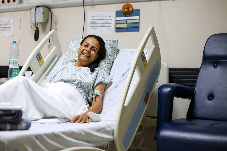 Uma mulher está deitada em uma cama de hospital， sorrindo. Ela usa um pijama hospitalar e tem um acesso venoso no braço. Ao lado da cama， há uma cadeira azul e uma garrafa de água. O ambiente é de um quarto de hospital， com paredes claras e alguns avisos na parede ao fundo.