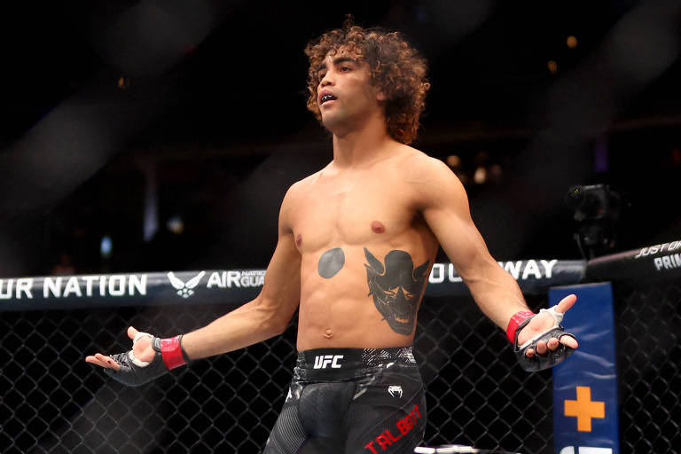 Um lutador de MMA está em uma arena， com os braços abertos e uma expressão de confiança. Ele está sem camisa， mostrando um tatuagem no peito. O ambiente é de uma luta de UFC， com uma grade ao fundo e iluminação focada no lutador.