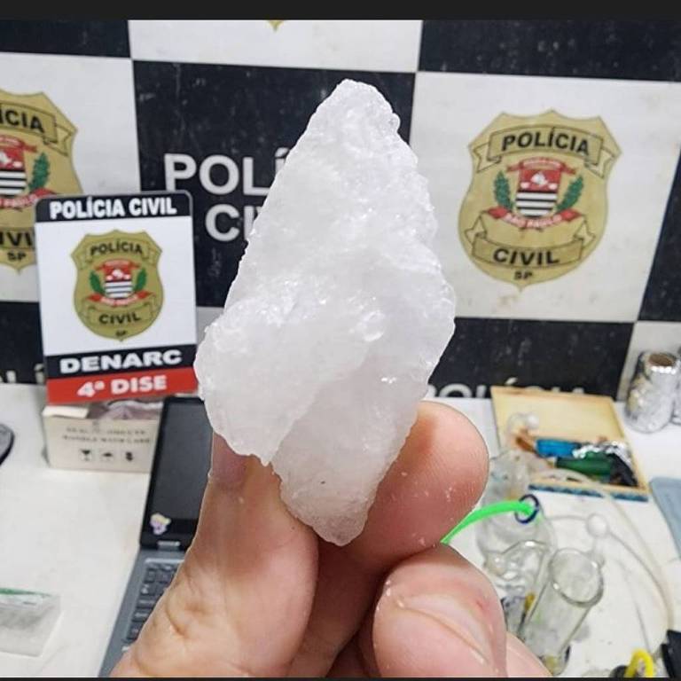 A imagem mostra uma mão segurando um cristal branco， que parece ser uma pedra de grande tamanho. Ao fundo， há um painel com emblemas da Polícia Civil e outros objetos， incluindo placas e documentos.