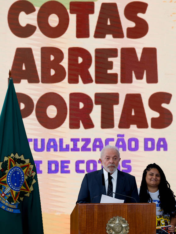 A imagem mostra um evento onde um homem， vestido com um terno escuro， está em pé ao lado de uma mulher com cabelo cacheado e vestido colorido. Eles estão em frente a um painel com as palavras 039;COTAS ABREM PORTAS039; em destaque， e abaixo， 039;ATUALIZAÇÃO DA LEI DE COTAS039;. À esquerda， há uma bandeira do Brasil. O ambiente parece ser formal， possivelmente uma conferência ou apresentação.