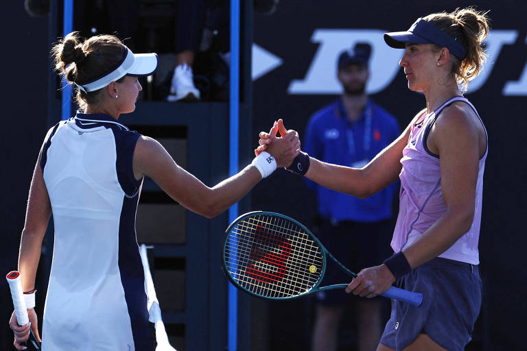 Duas jogadoras de tênis estão se cumprimentando após uma partida. A jogadora à esquerda usa uma camiseta branca com detalhes em azul e uma faixa na cabeça， enquanto a jogadora à direita veste uma camiseta roxa e shorts escuros. Ambas seguram raquetes de tênis. Ao fundo， um homem está sentado， observando.
