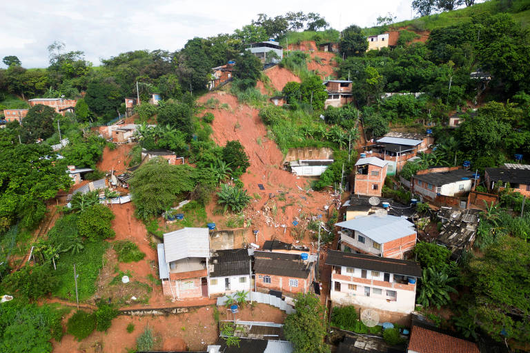 A imagem mostra uma área urbana afetada por um deslizamento de terra. Várias casas estão localizadas em uma encosta， com algumas delas parcialmente cobertas por terra e vegetação. O terreno é verde， com árvores e arbustos ao redor， e o céu está nublado.
