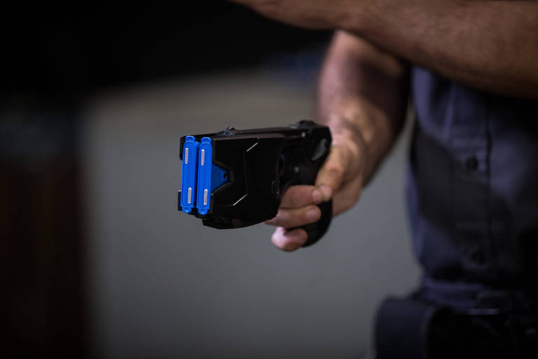 A imagem mostra uma mão segurando uma arma de taser， que é preta com detalhes azuis. O fundo é desfocado， sugerindo um ambiente interno.
