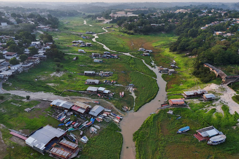 A imagem mostra uma vista aérea de uma área rural com um leito de rio seco e vegetação ao redor. Há várias construções simples， algumas de metal e outras de madeira， dispostas ao longo do rio. O terreno é predominantemente verde， com algumas áreas de solo exposto. Ao fundo， é possível ver uma área mais urbanizada com edifícios e árvores.
