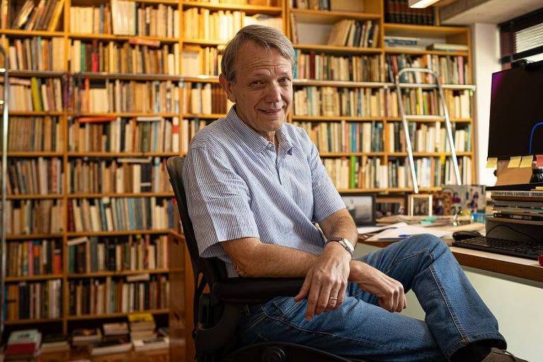 Um homem sorridente está sentado em uma cadeira em frente a uma mesa, com uma estante cheia de livros ao fundo. Ele usa uma camisa de gola clara e jeans. A mesa tem alguns livros e um computador. O ambiente é bem iluminado, com uma atmosfera acolhedora.