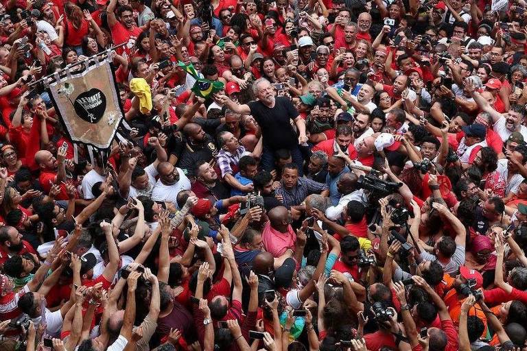 A imagem mostra uma grande multidão vestida predominantemente de vermelho, reunida em um evento público. Algumas pessoas estão levantando as mãos, enquanto outras seguram uma bandeira ou cartaz. No centro, uma figura é erguida por membros da multidão, que parecem estar celebrando ou demonstrando apoio.