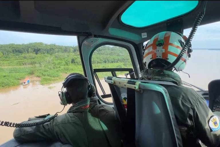 A imagem mostra o interior de um helicóptero， com dois pilotos visíveis. Um deles está usando um capacete com listras laranjas e fones de ouvido， enquanto o outro está de perfil， também com fones. A vista externa revela uma área verde e um corpo d039;água abaixo.