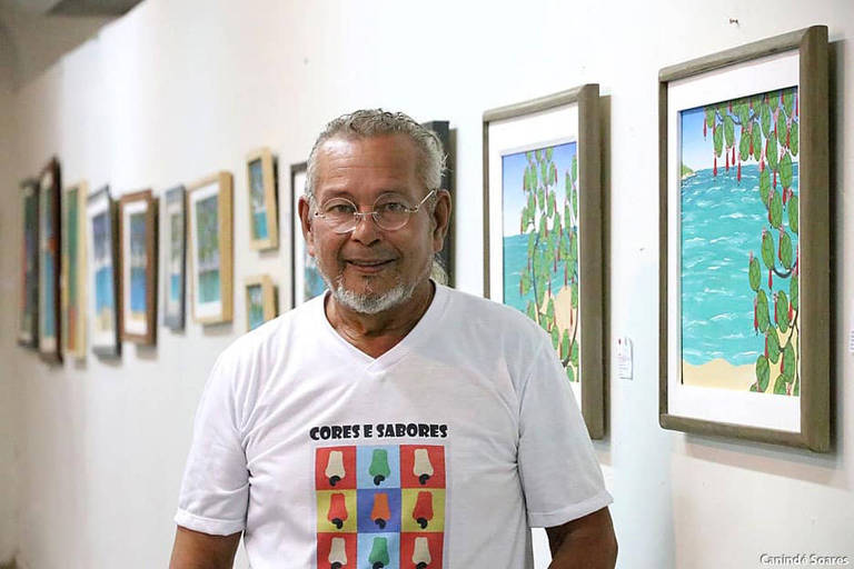 Um homem sorridente está posando em uma galeria de arte， com várias obras emolduradas ao fundo. Ele usa uma camiseta branca com uma estampa colorida. As obras na parede apresentam paisagens e elementos naturais.