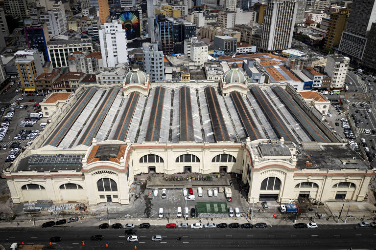 Imagem aérea de um grande mercado com um telhado de várias seções. O edifício é de estilo arquitetônico clássico， com janelas grandes e arcos. Ao redor， há uma densa área urbana com prédios altos e estacionamentos visíveis.