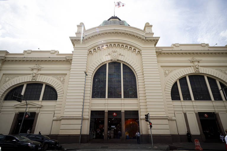 A imagem mostra uma torra na fachada do Mercado Municipal de São Paulo, um edifício de estilo arquitetônico neoclássico. A estrutura é predominantemente branca, com grandes janelas em arco e detalhes ornamentais. No topo, é possível ver parte da cúpula da torre e uma bandeira do município de São Paulo. A entrada principal é ampla, com portões de ferro abertos. Na parte inferior, há sinais de trânsito e pessoas caminhando na calçada.